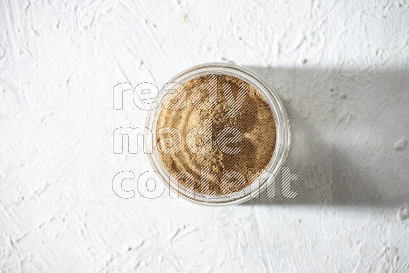 A glass jar full of cumin powder on textured white flooring