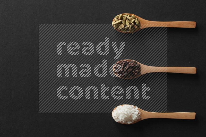 wooden spoons filled with white peppers, cloves, cardamom, salt, black peppers and basil on black flooring