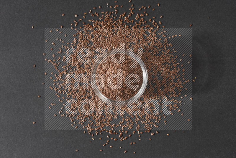 وعاء زجاجي ممتلئ بحبوب بذر الكتان محاط بالبذور على أرضية سوداء