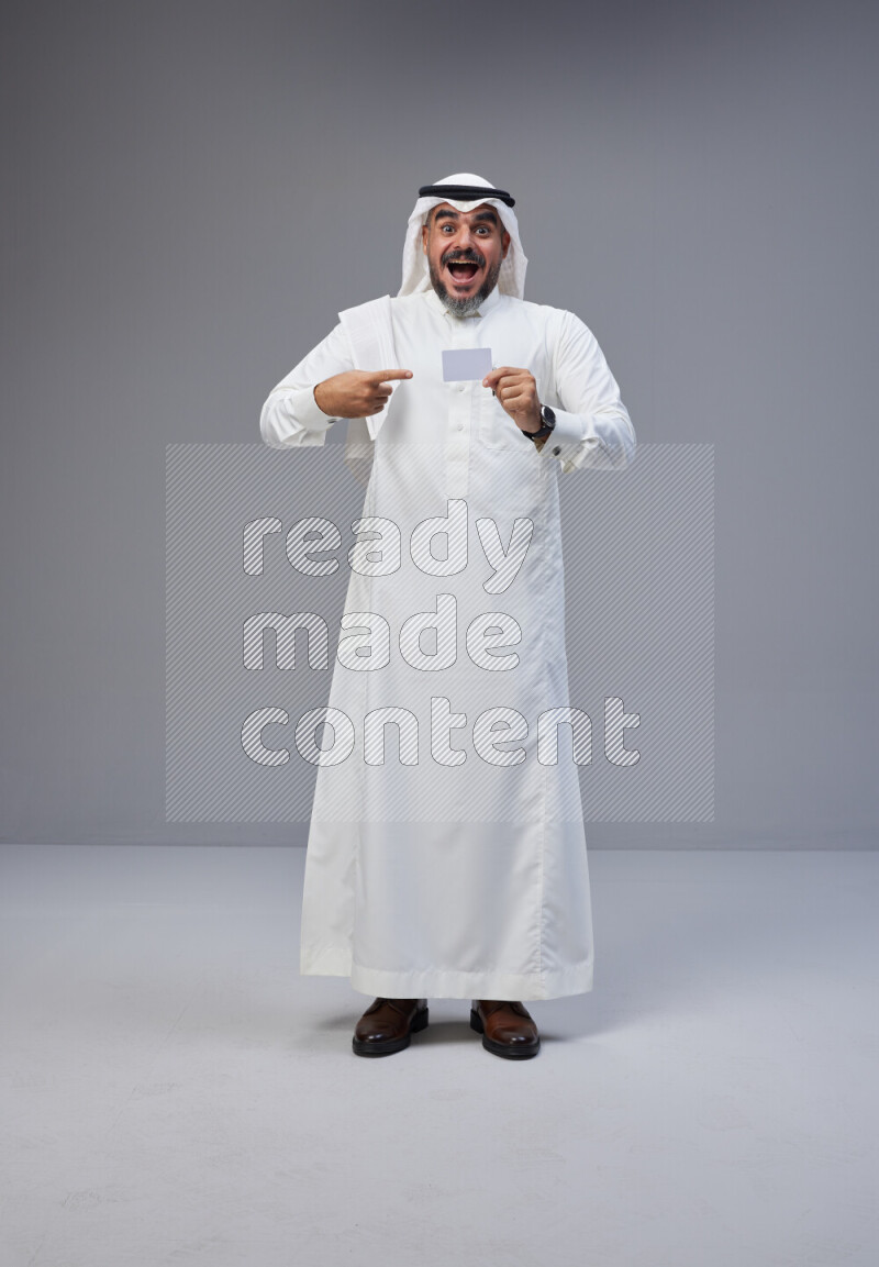 Saudi man Wearing Thob and white Shomag standing holding ATM card on Gray background