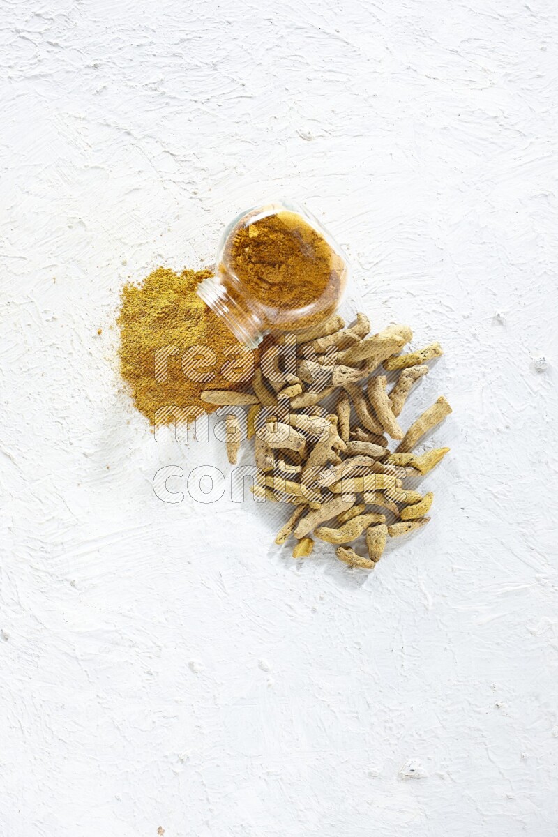 A flipped glass spice jar full of turmeric powder and powder spilled out of it with dried whole fingers on textured white flooring