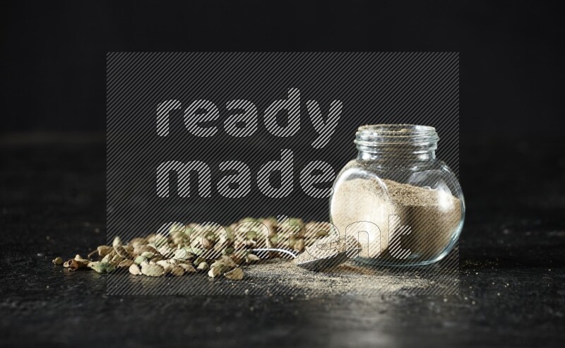 وعاء زجاجي للتوابل وملعقة معدنية ممتلئة ببودرة الهيل وحبوب الهيل متناثرة على خلفية سوداء