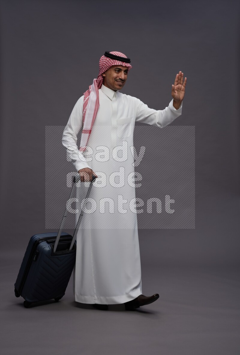 Saudi man wearing thob and shomag standing holding bag on gray background