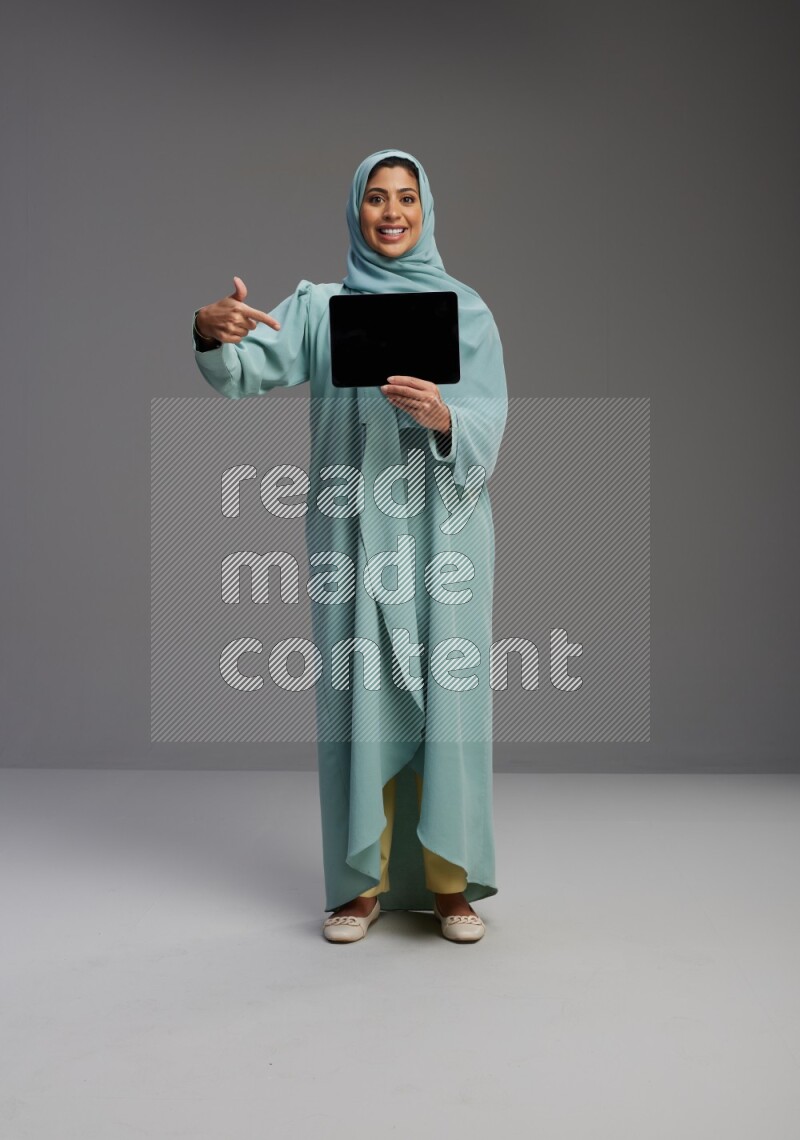Saudi Woman wearing Abaya standing showing tablet to camera on Gray background