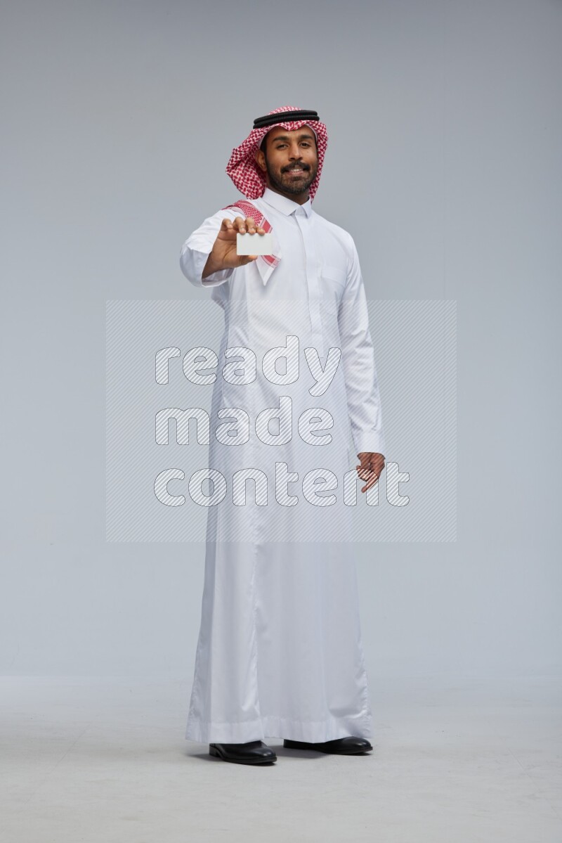 Saudi man wearing thob and shomag standing holding ATM card on gray background