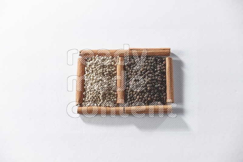 مربعان من أعواد القرفة ممتلئان بحبوب الفلفل الأسود والفلفل الأبيض علي خلفية بيضاء