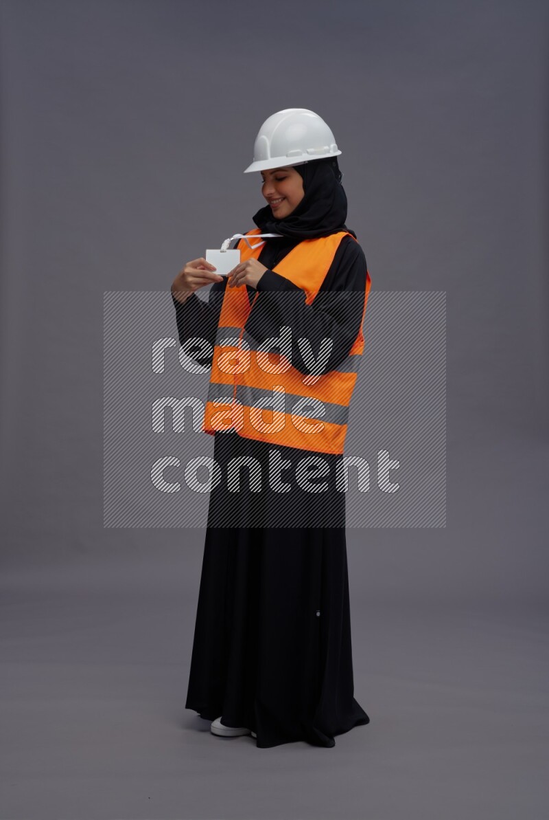 Saudi woman wearing Abaya with engineer vest with neck strap employee badge standing interacting with the camera on gray background
