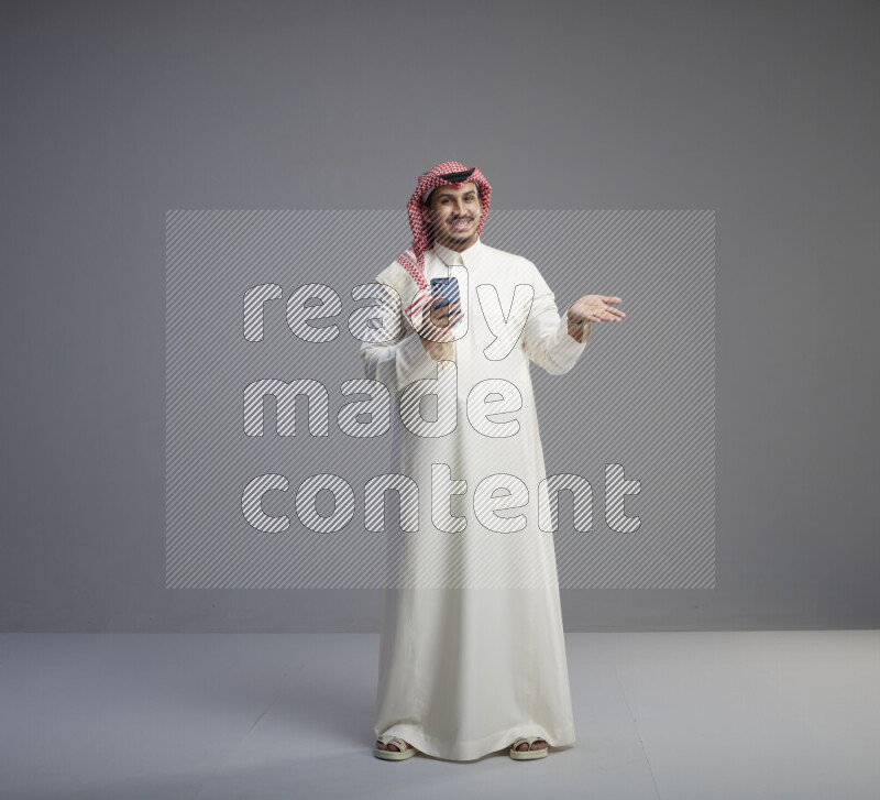 A  Saudi man standing wearing thob and red shomag texting on phone on gray background