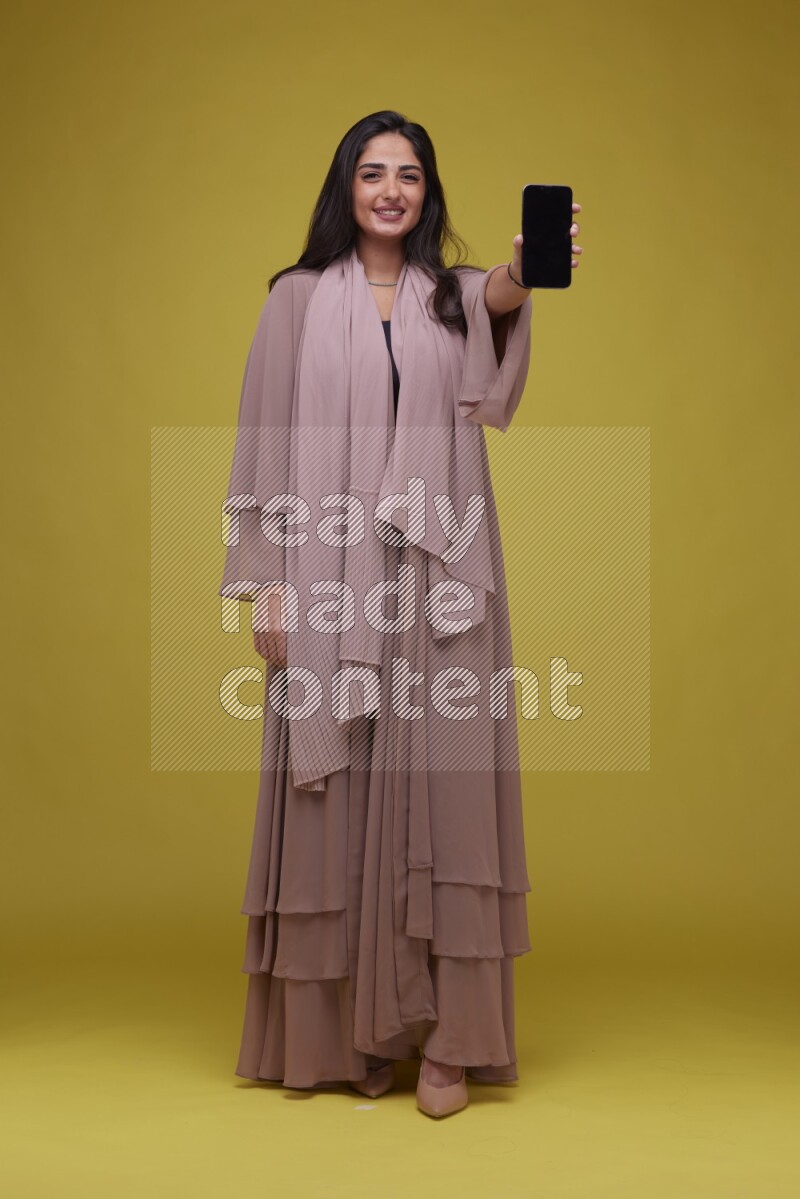 A woman Showing SmartPhone Screen on a Yellow Background wearing Brown Abaya