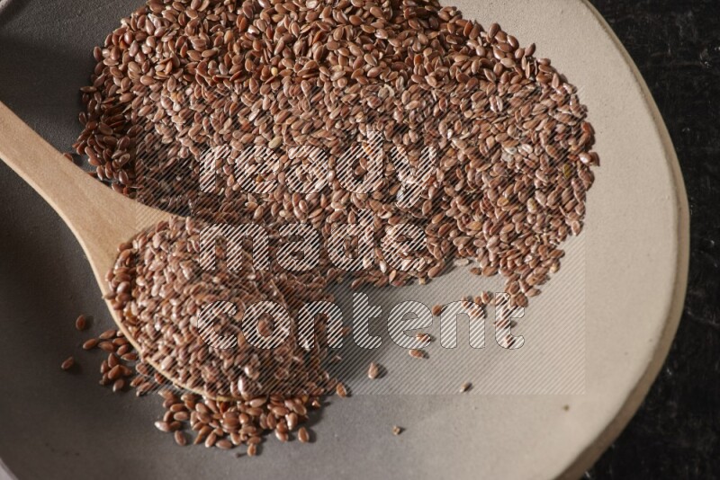 A multicolored pottery plate full of flaxseeds and wooden spoon full of seeds on a textured black flooring