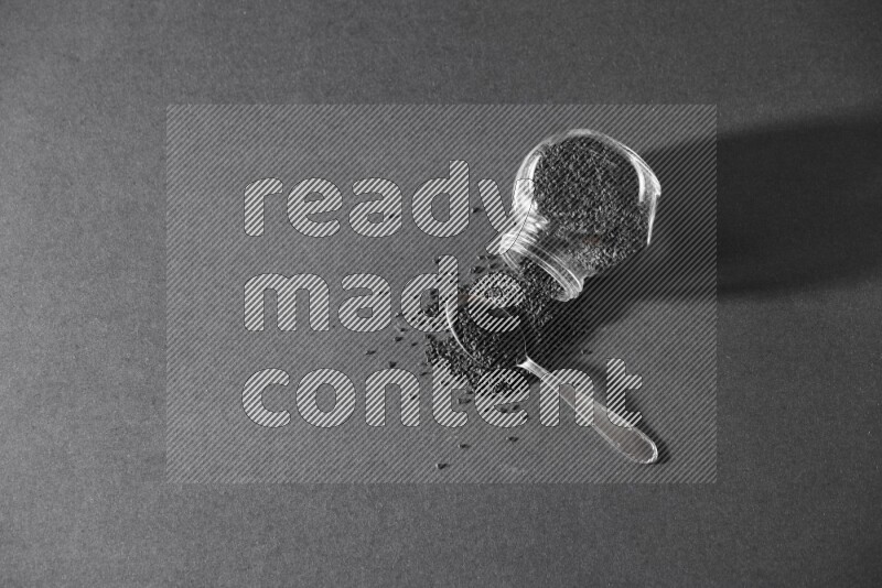 A flipped glass spice jar full of black seeds and the seeds spread out with a metal spoon full it on a black flooring