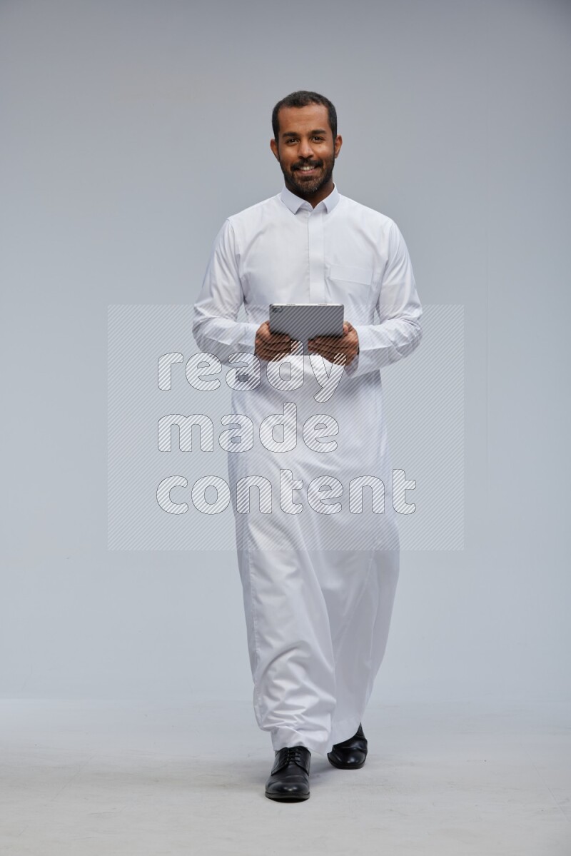 Saudi man wearing Thob standing working on tablet on Gray background