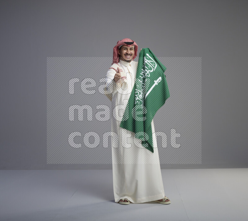 A saudi man standing wearing thob and red shomag holding big saudi flag on gray background