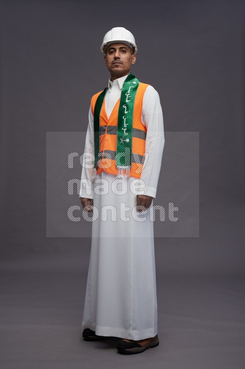Saudi man wearing thob with engineer vest standing holding Saudi flag on gray background