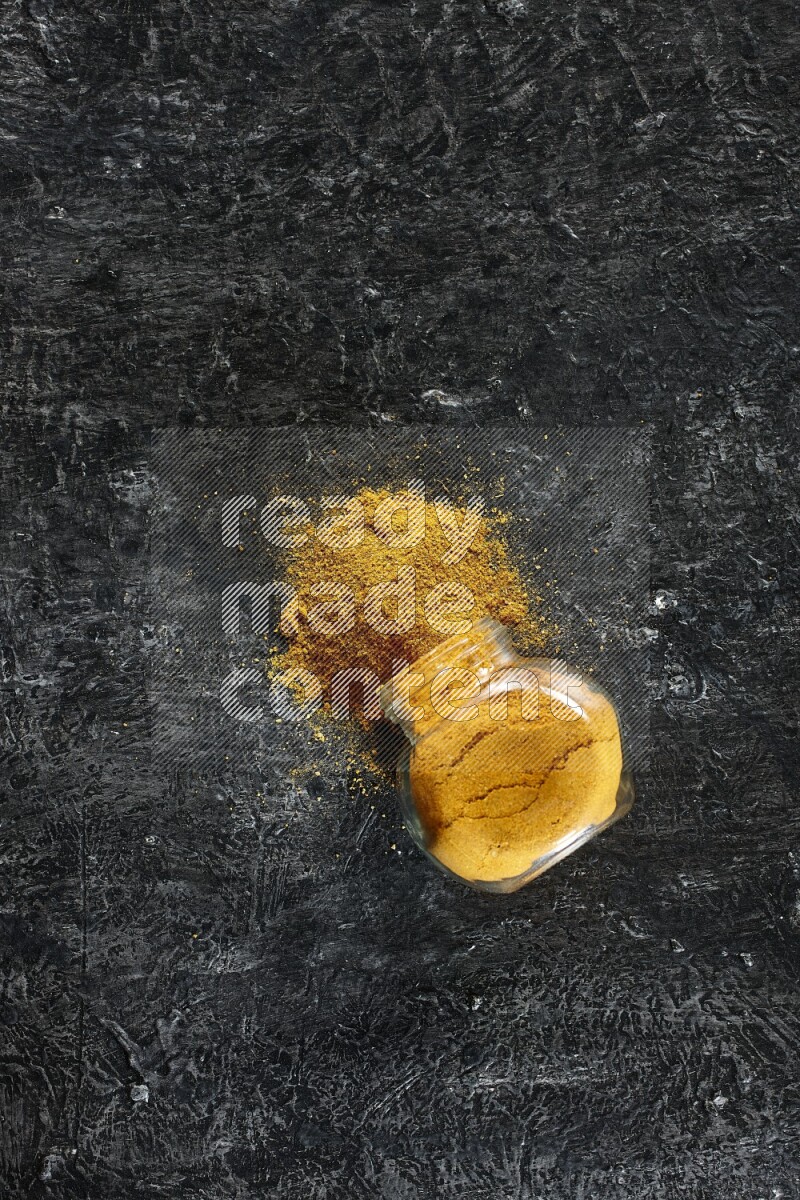 A flipped glass spice jar full of turmeric powder and powder spilled out of it on textured black flooring