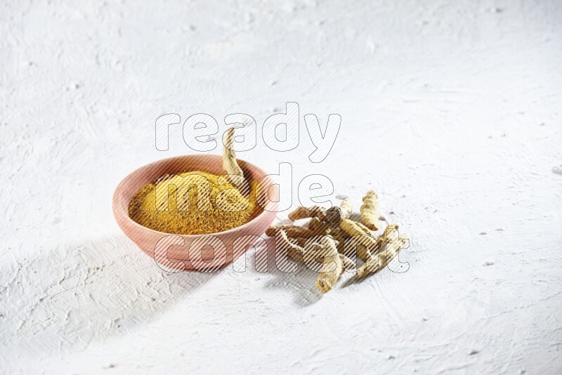 A wooden bowl full of turmeric powder and dried turmeric whole fingers beside it on textured white flooring
