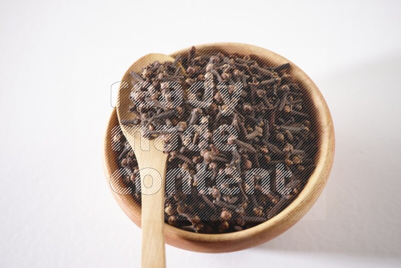 A wooden bowl and a wooden spoon full of cloves on a white flooring