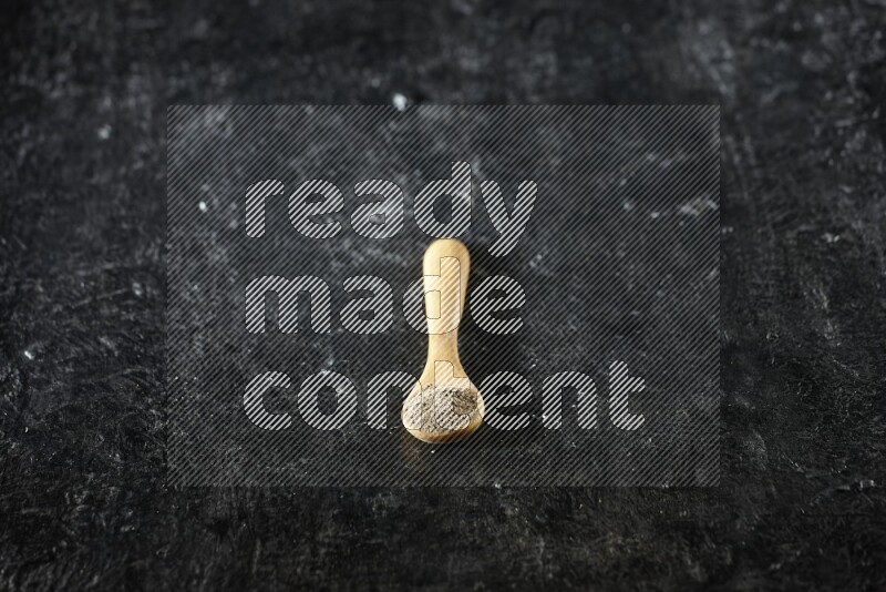 A wooden spoon full of cardamom powder on textured black flooring