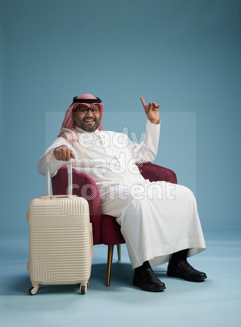 Saudi Man with shimag sitting on chair with a travel luggage on blue background