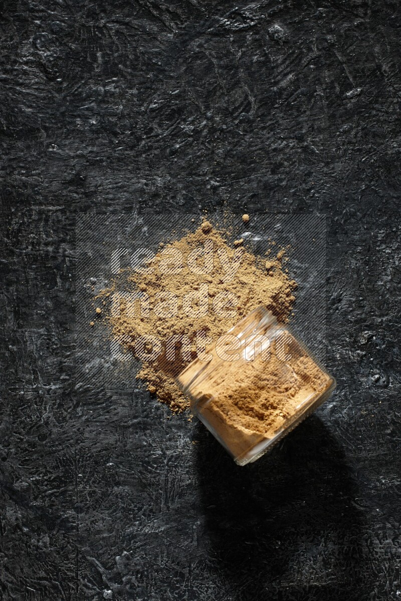 A flipped glass jar full of allspice powder and powder spilled out of it on a textured black flooring