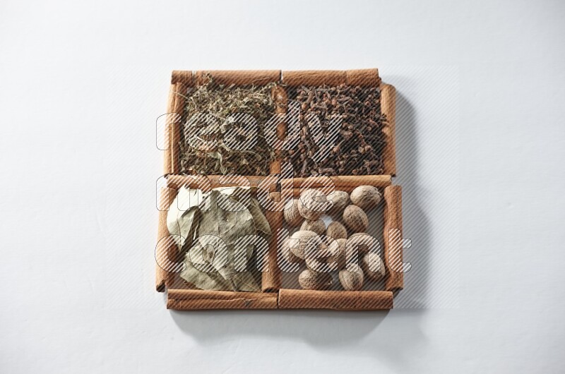 4 مربعات من أعواد القرفة ممتلئة بأوراق الغار والريحان المجفف والقرنفل وجوزة الطيب على أرضية بيضاء