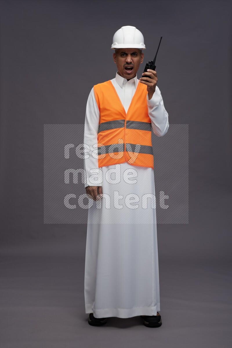 Saudi man wearing thob with engineer vest standing holding walkie-talkie on gray background