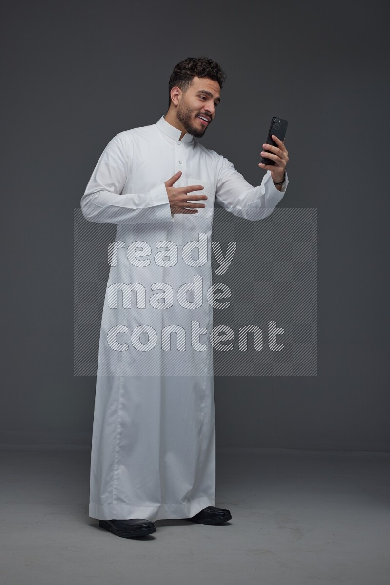 A Saudi man wearing Thobe and taking selfie with his phone making different poses eye level on a gray background