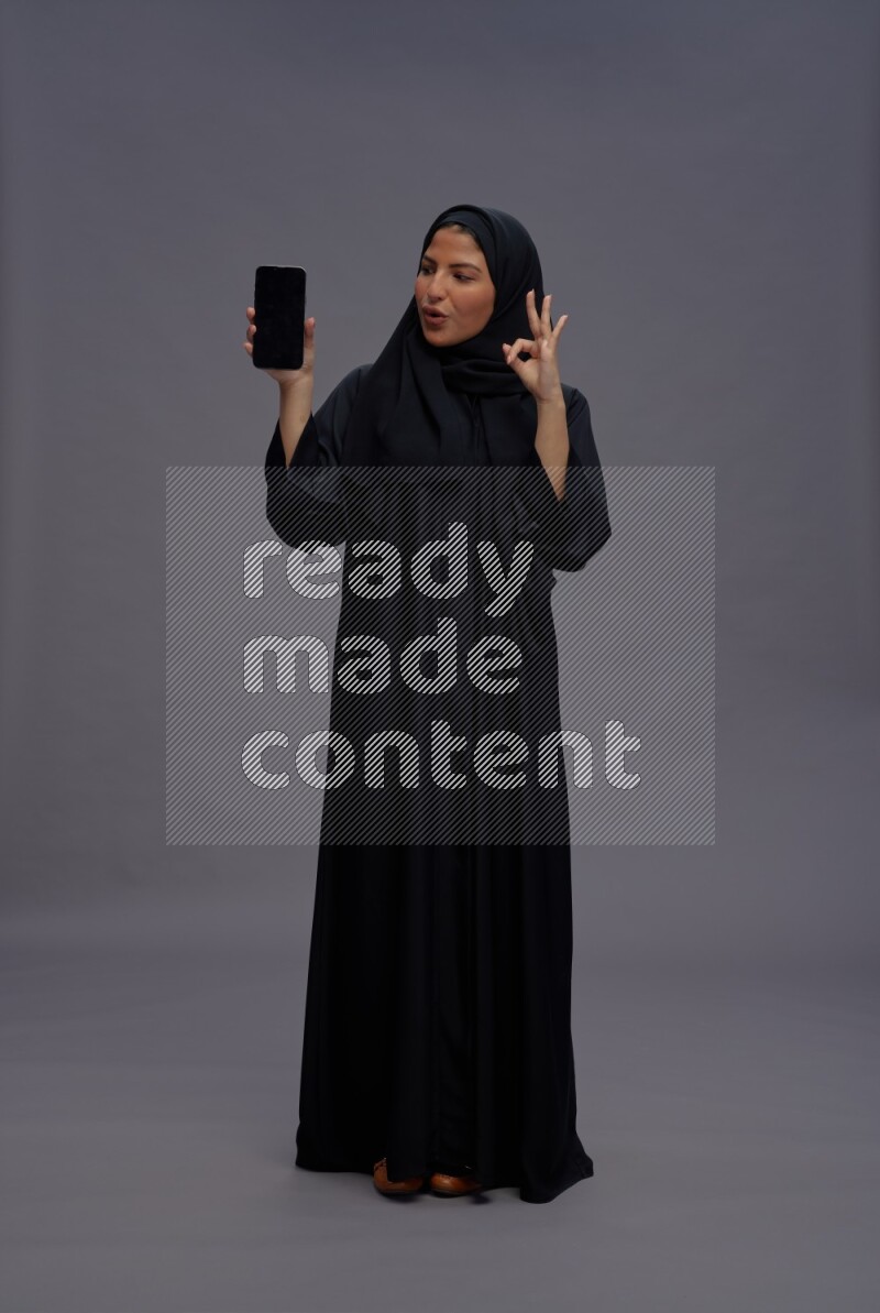 Saudi woman wearing Abaya standing showing phone to camera on gray background