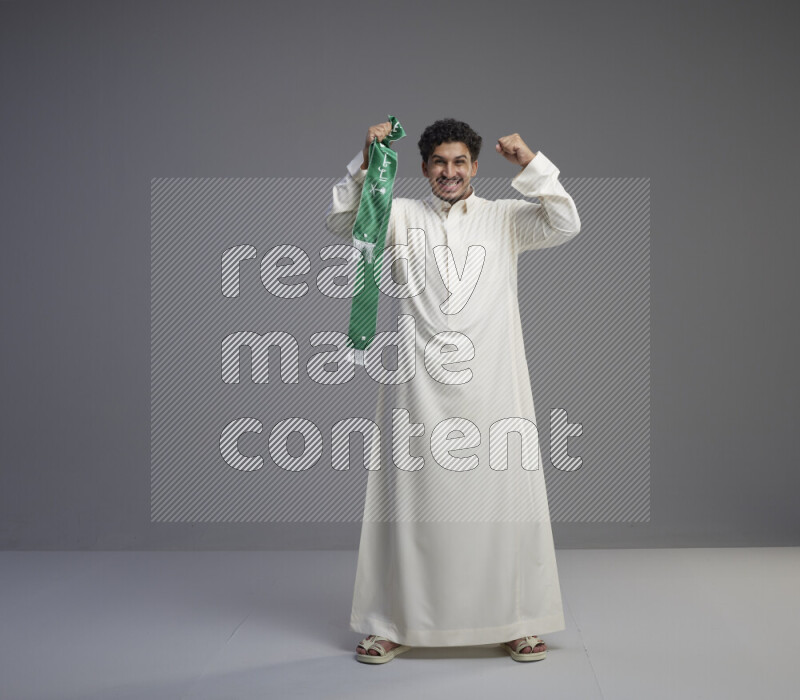 A Saudi man standing wearing thob holding Saudi flag scarf on gray background