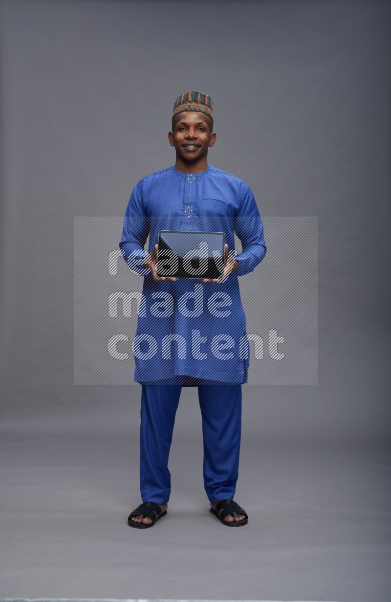 Man wearing Nigerian outfit standing showing tablet to camera on gray background