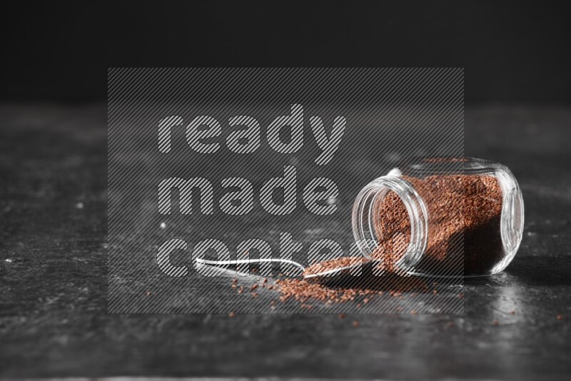 وعاء زجاجي مقلوب ممتلئ ببذور حب الرشاد مع بعض الحبوب المتناثرة منه مع ملعقة معدنية ممتلئة بالبذور على أرضية سوداء