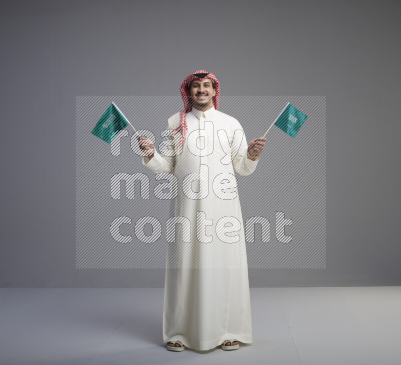A Saudi man standing wearing thob and red shomag raising small Saudi flag on gray background