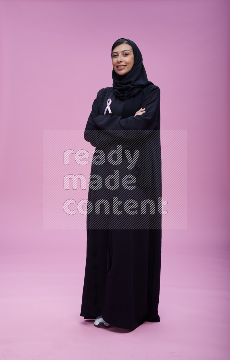 Saudi woman wearing pink ribbon on Abaya standing crossed arms on pink background