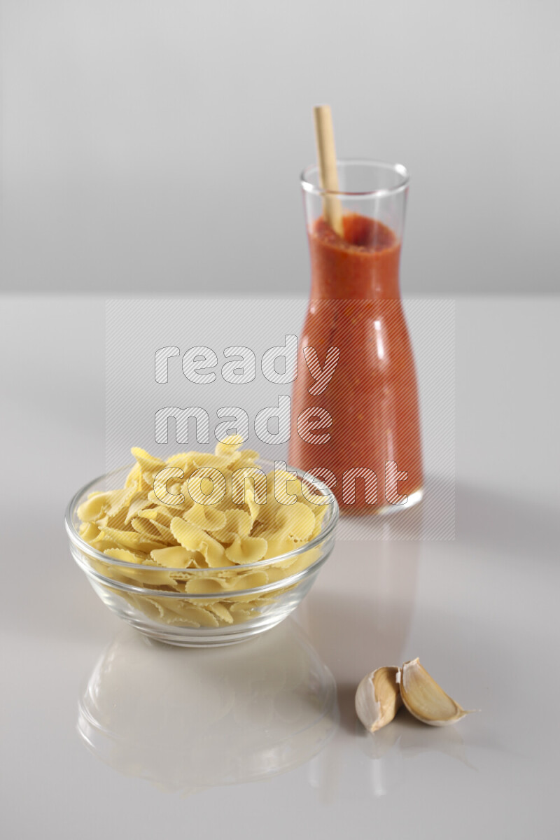 Raw pasta with tomatoe pasta with different ingredients such as cherry tomatoes, basil, garlic, bay laurel, cardamom, white pepper, black pepper, red chilis and wheat stalks on light grey background