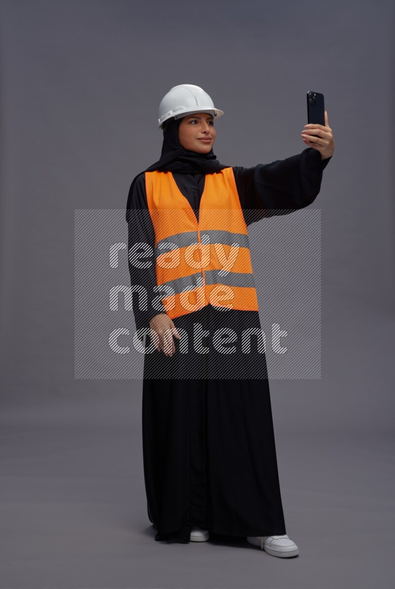 Saudi woman wearing Abaya with engineer vest standing taking selfie on gray background