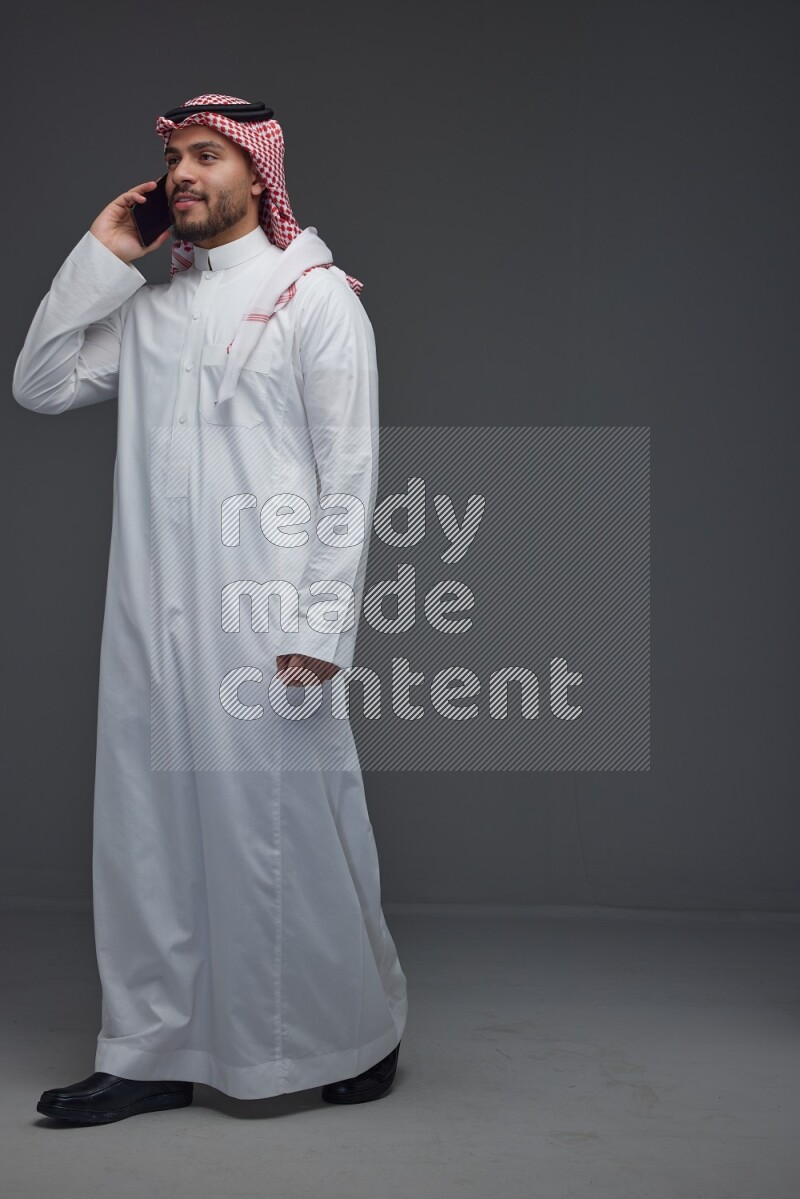 A Saudi man wearing Thobe and Shmagh talking in the phone eye level on a gray background