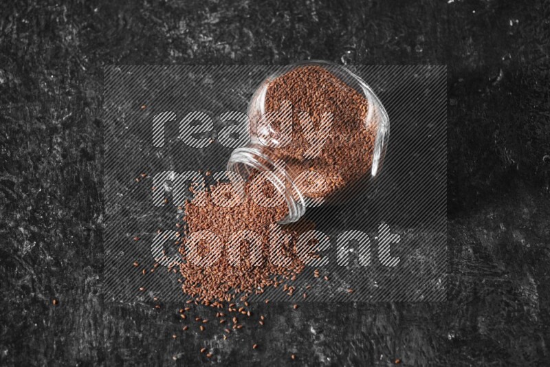 وعاء زجاجي مقلوب ممتلئ ببذور حب الرشاد وتناثرت البذور منه على أرضية سوداء