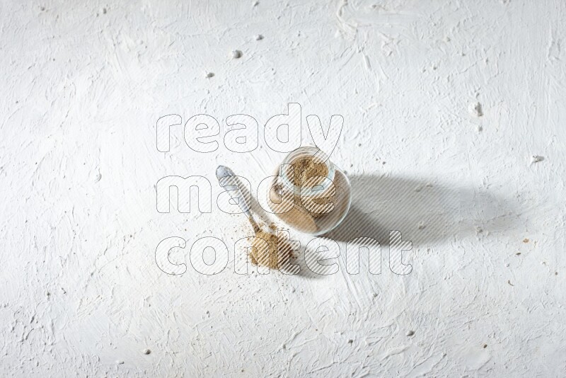 A glass spice jar and metal spoon full of cumin powder on textured white flooring
