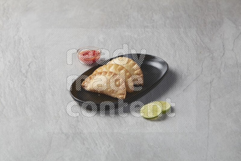 Three fried sambosas in an oval shaped black plate and a red sauce in a black round ramekin on a gray background