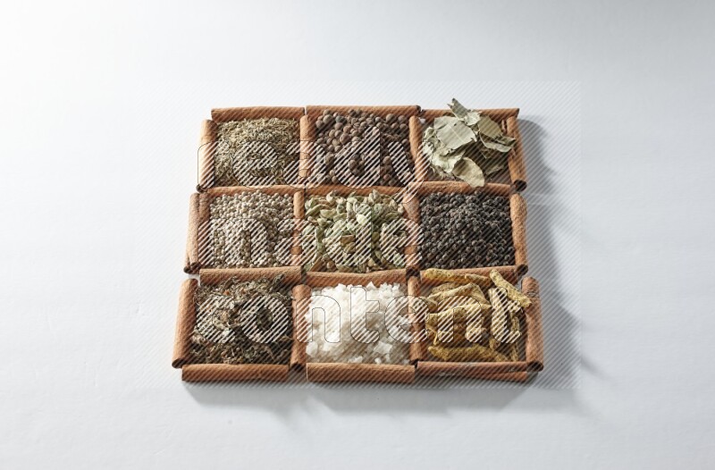 9 squares of cinnamon sticks full of cardamom in the middle surrounded by turmeric, salt, dried basil, white pepper, cumin, allspice, bay laurel leaves and black pepper on white flooring