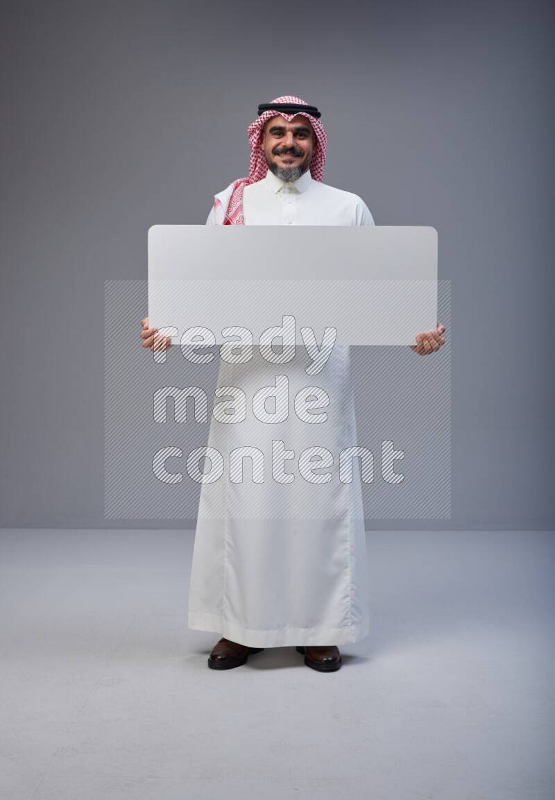 Saudi man Wearing Thob and red Shomag standing holding board on Gray background