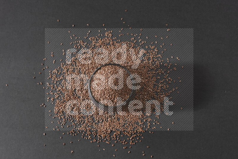 A black pottery bowl full of flaxseeds surrounded by the seeds on a black flooring