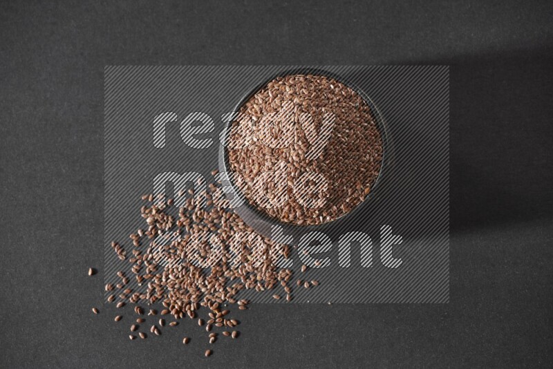 A black pottery bowl full of flaxseeds surrounded by the seeds on a black flooring