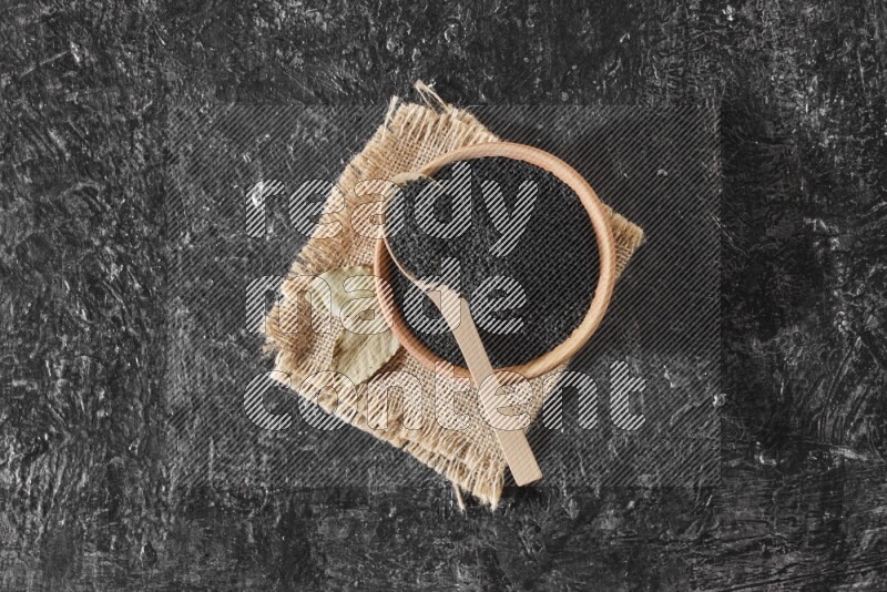 A wooden bowl full of black seeds with wooden spoon full of the seeds on it on a burlap fabric on a textured black flooring
