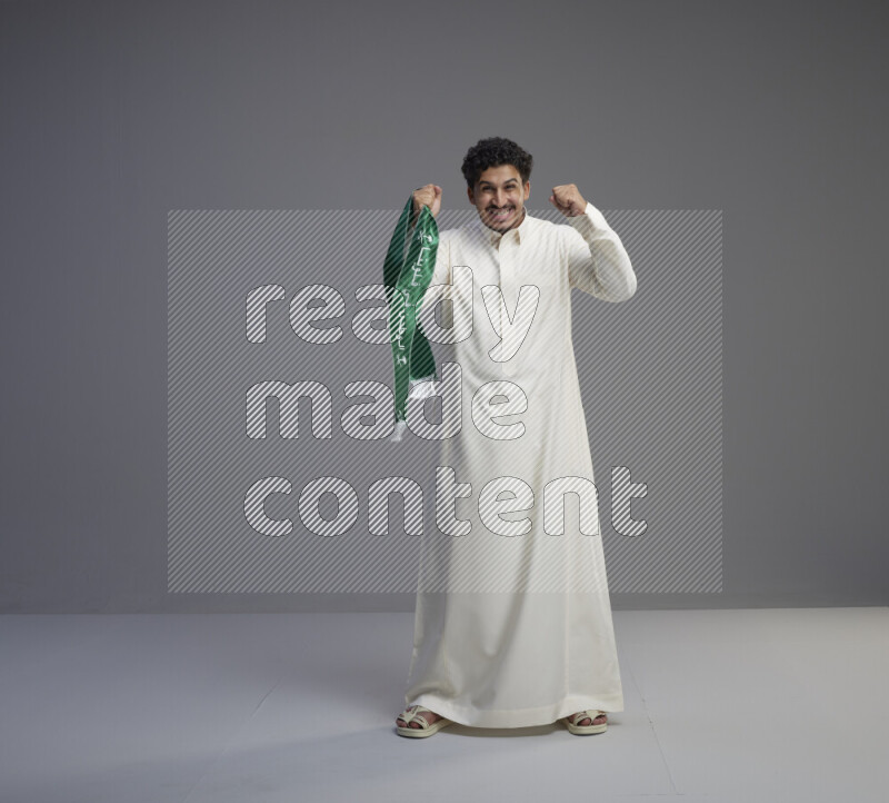 A Saudi man standing wearing thob holding Saudi flag scarf on gray background