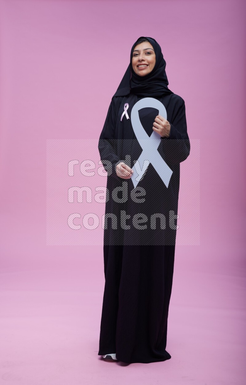 Saudi woman wearing pink ribbon on abaya standing holding awareness ribbon on plain pink background