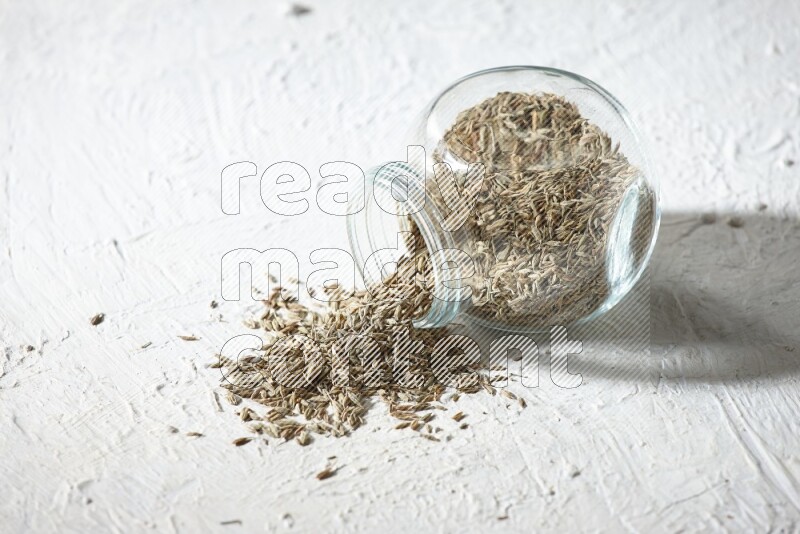 A flipped glass spice jar full of cumin seeds and the seeds spilled out on textured white flooring