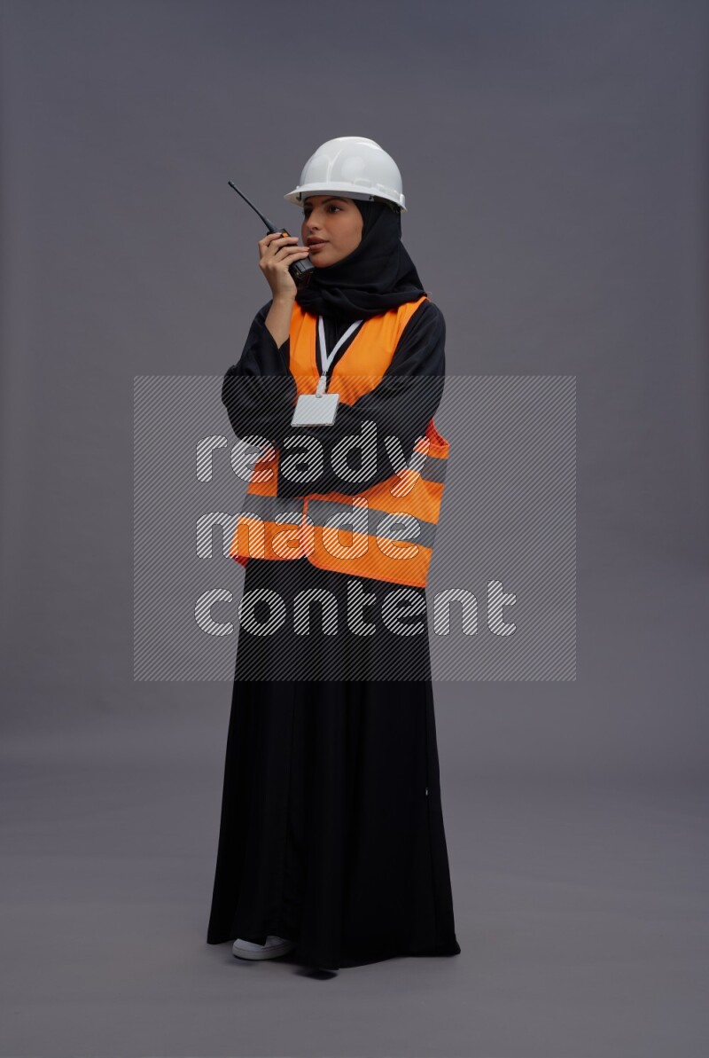 Saudi woman wearing Abaya with engineer vest with neck strap employee badge standing holding walkie-talkie on gray background
