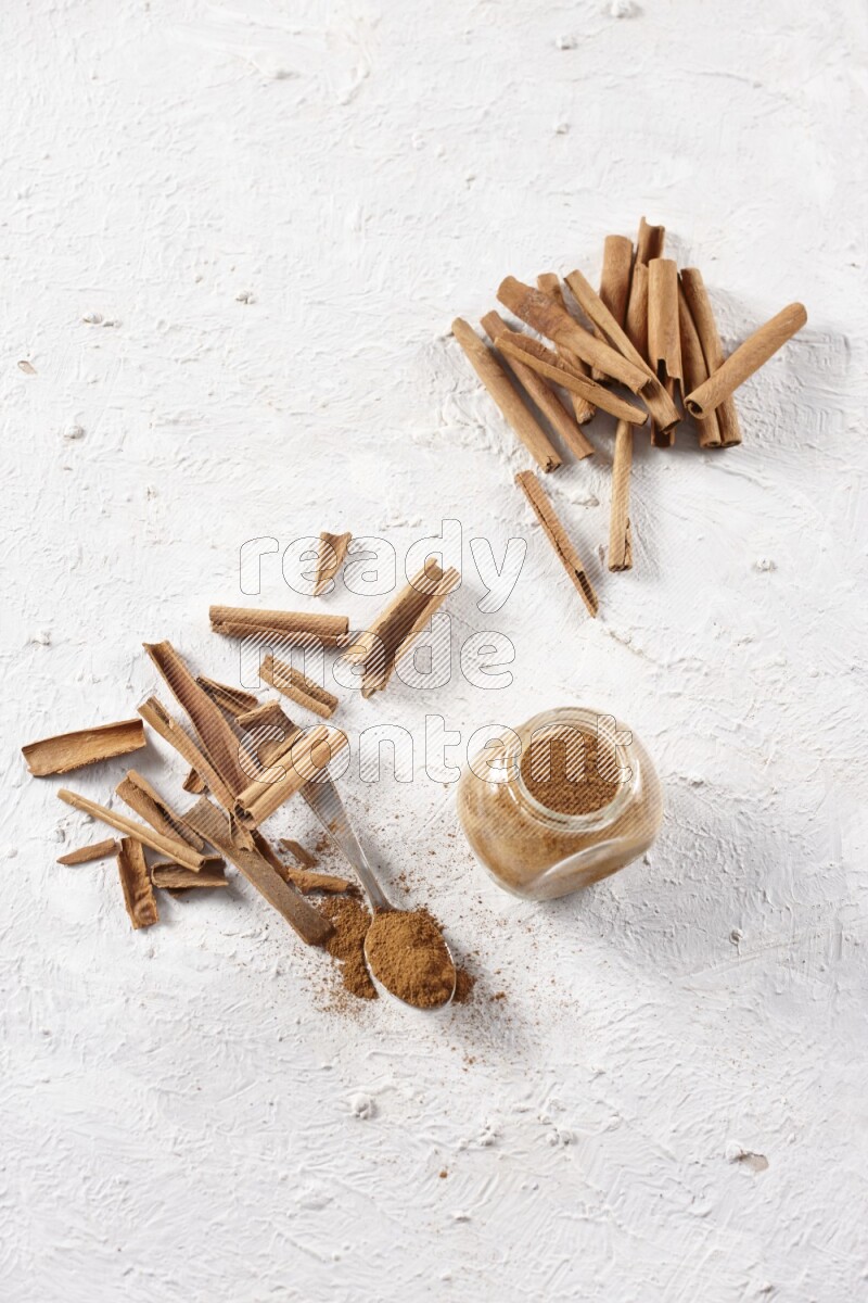 Herbal glass jar full cinnamon powder and a metal spoon surrounded by cinnamon sticks on a white background