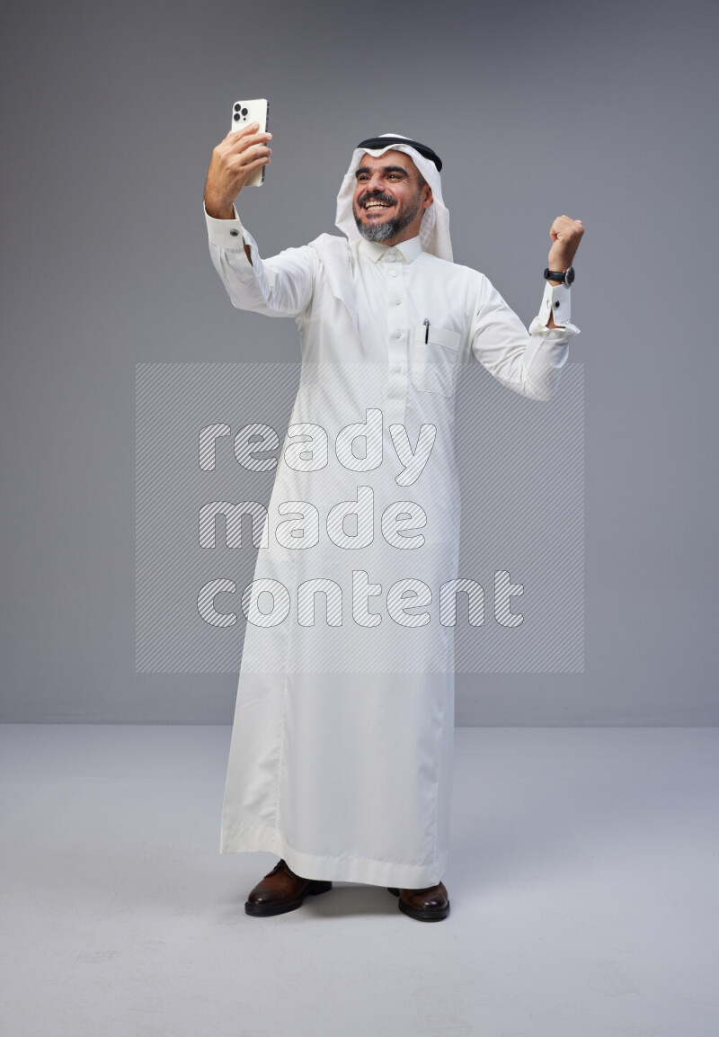 Saudi man Wearing Thob and white Shomag standing taking selfie on Gray background